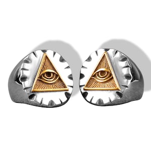 Op de afbeelding: Twee zilverkleurige ringen met een cirkelvormig voorvlak met een goudkleurige inleg van een driehoek met een oog. De zilveren band heeft een getextureerde, ingekeepte rand rondom het centrale goudkleurige piramide- en oogontwerp op elke ring.