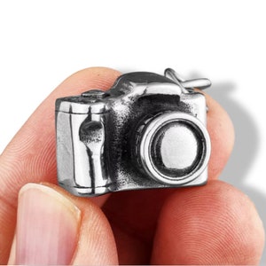 May include: A silver-colored camera pendant with a detailed design. The pendant features a lens, buttons, and other camera details. The camera is held in a hand against a white background.