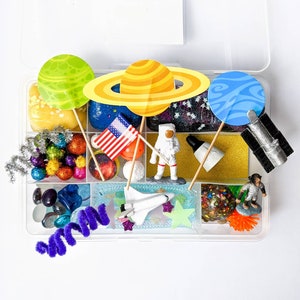 May include: A clear plastic storage box filled with various space-themed toys and craft supplies. The box includes planets, an astronaut, a spaceship, a flag, and colorful beads.