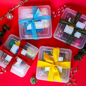 May include: Four clear plastic boxes tied with colorful ribbons, each containing various items. The boxes are arranged on a red surface, with holiday decorations scattered around. The boxes have a label that says "Busy Hands Through Play".