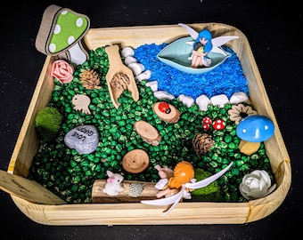 May include: A wooden tray filled with green beans, a blue river, white rocks, and various fairy garden accessories, including a mushroom, a ladybug, a flower, and two fairies. The tray also includes a gray heart-shaped stone with the words "Love Garden" written on it.