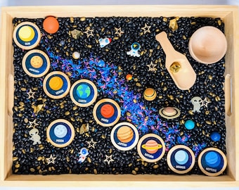 May include: A wooden tray filled with black beans, gold stars, and blue and purple rice.  There are nine wooden circles with planets and their names: Sun, Mercury, Venus, Earth, Mars, Jupiter, Saturn, Uranus, and Neptune.  There are also small wooden alien figures and a wooden scoop.
