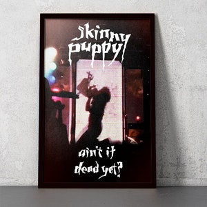 May include: A black and white poster with the band name "Skinny Puppy" in white lettering. The poster features a silhouette of a person in front of a screen with the words "ain't it dead yet?" in white lettering.