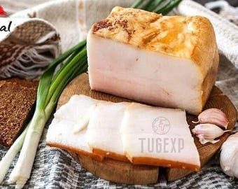 May include: A close-up of a wooden cutting board with slices of white, cured pork fat, also known as lard, with a few cloves of garlic and green onions. The board is on a white and brown patterned fabric.