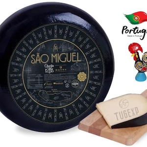 May include: A round wheel of São Miguel cheese wrapped in black wax with a gold label. The label reads "Queijo Ilha Velho 9 Meses" and features a map of the island of São Miguel. The cheese is on a wooden cutting board with a wedge of cheese cut off.