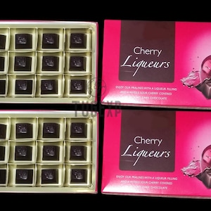 Op de afbeelding: Twee dozen met chocolade-kersenlikeur-pralines. Elke doos bevat 24 individueel verpakte pralines. De dozen zijn roze met witte tekst die "Cherry Liqueurs" luidt. De dozen zijn versierd met een kersen- en chocoladeontwerp.