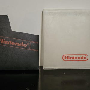 以下が含まれることがあります： 赤い Nintendo ロゴがついた黒い Nintendo ゲームカートリッジ。カートリッジは、赤い Nintendo ロゴがついた透明なプラスチックケースの隣にあります。
