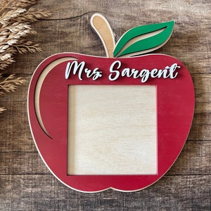 May include: A wooden apple-shaped frame with a red exterior and a natural wood interior. The frame has a green leaf and a tan stem. The name "Mrs. Sargent" is written in white script across the top. A square opening is in the center.