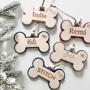 May include: Five bone-shaped wooden ornaments with colored backings, each featuring a name, a year, and small icons like paw prints or angel wings. The ornaments are suspended by twine with wooden beads and include the names Indie, Remi, Kali, Bella, and STITCH.
