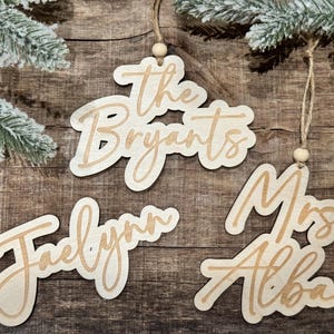 May include: Four wooden ornaments with names written in cursive. The ornaments are "The Bryants", "Jaelynn", "Mrs. Alba", and are all hanging from twine.