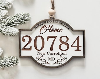 May include: A decorative wooden hanging ornament with a white center and dark brown frame. It features the text "THERE'S NO PLACE LIKE Home", the number "20784", "New Carrollton", and "MD" with a small branch graphic, suspended by a twine cord.