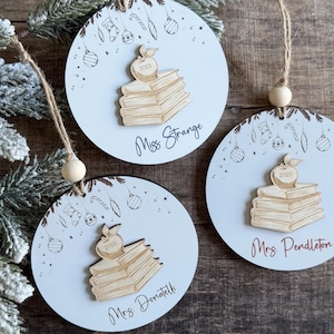 May include: Three round, light blue Christmas ornaments with a teacher theme. Each ornament features a stack of books with an apple on top, and the teacher's name. The ornaments are hanging from twine.