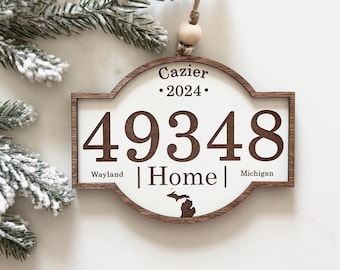 May include: A wooden ornament with a white center featuring the text Cazier 2024, the number 49348, the word Home, and the text Wayland Michigan. A small brown silhouette of the state of Michigan is printed at the bottom. It hangs from a twine loop.