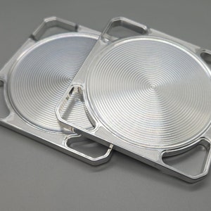 May include: Two silver metal coasters with a circular, concentric design. Each coaster has four square cutouts around the edge.