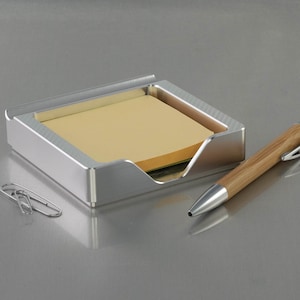Peut inclure: Un porte-notes adhésifs en métal argenté avec un bloc-notes jaune à l'intérieur et un stylo en bois avec un clip argenté sur une surface argentée.