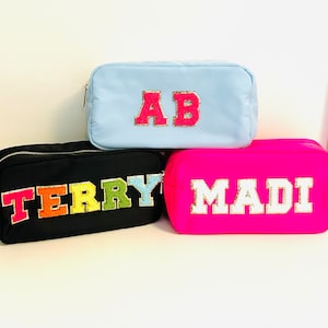May include: Three different cosmetic bags with personalized names. The top bag is light blue with the name "AB" in pink and gold letters. The middle bag is black with the name "TERRY" in rainbow letters. The bottom bag is bright pink with the name "MADI" in white and gold letters.