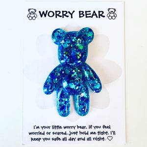 Op de afbeelding: Blauwe glitter Worry Bear-beeldje op een witte kaart. Het beer-vormige beeldje heeft een sprankelend, getextureerd uiterlijk. De kaart heeft bovenaan de woorden "WORRY BEAR" en onderaan een bericht.