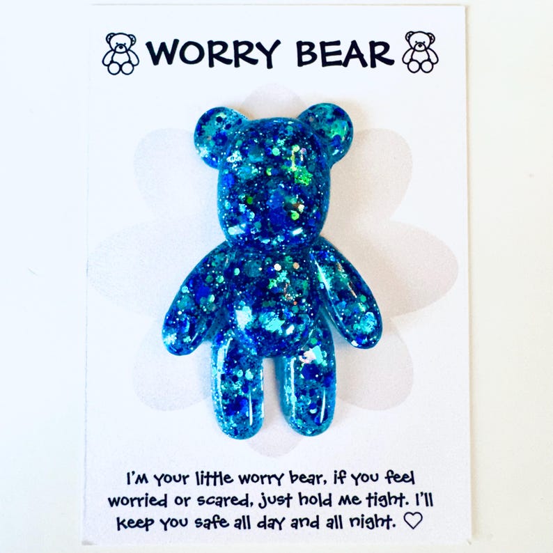 Op de afbeelding: Blauwe glitter Worry Bear-beeldje op een witte kaart. Het beer-vormige beeldje heeft een sprankelend, getextureerd uiterlijk. De kaart heeft bovenaan de woorden "WORRY BEAR" en onderaan een bericht.