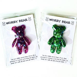 Op de afbeelding: Twee glitterhars worry bears, &eacute;&eacute;n paars en &eacute;&eacute;n groen, verpakt op kaarten. Elke beer heeft de vorm van een teddybeer en is bedekt met glitter. De kaarten lezen "Worry Bear" en bevatten een geruststellende boodschap.