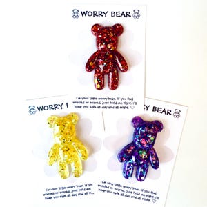 Op de afbeelding: Drie glitterende Worry Bear beeldjes van hars in rood, geel en paars, elk op een kaart. De kaarten hebben de tekst "WORRY BEAR" en een bericht: "I'm your little worry bear...keep you safe all day and all night."