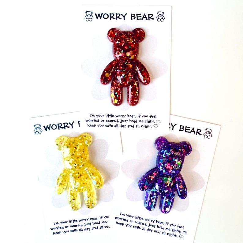 Op de afbeelding: Drie glitterende Worry Bear beeldjes van hars in rood, geel en paars, elk op een kaart. De kaarten hebben de tekst "WORRY BEAR" en een bericht: "I'm your little worry bear...keep you safe all day and all night."