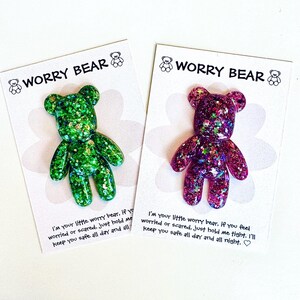 Op de afbeelding: Twee glitterende Worry Bear-beeldjes, &eacute;&eacute;n groen en &eacute;&eacute;n paars, elk op een kaart. De kaarten hebben de tekst "WORRY BEAR" en een boodschap over het bieden van troost. De beren zijn in een schattige, cartoonstijl.
