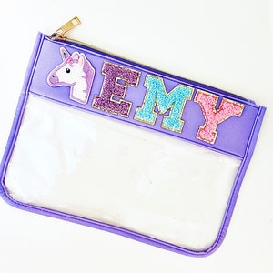 May include: Purple zippered pouch with a clear plastic window. The pouch has a unicorn and the name "EMY" in colorful embroidered letters.