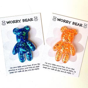 Op de afbeelding: Twee glitterhars Worry Bear-bedels, &eacute;&eacute;n blauw en &eacute;&eacute;n oranje, op witte kaarten. Elke kaart heeft de tekst "Worry Bear" en een geruststellende boodschap. De beren zijn gevormd als teddyberen.