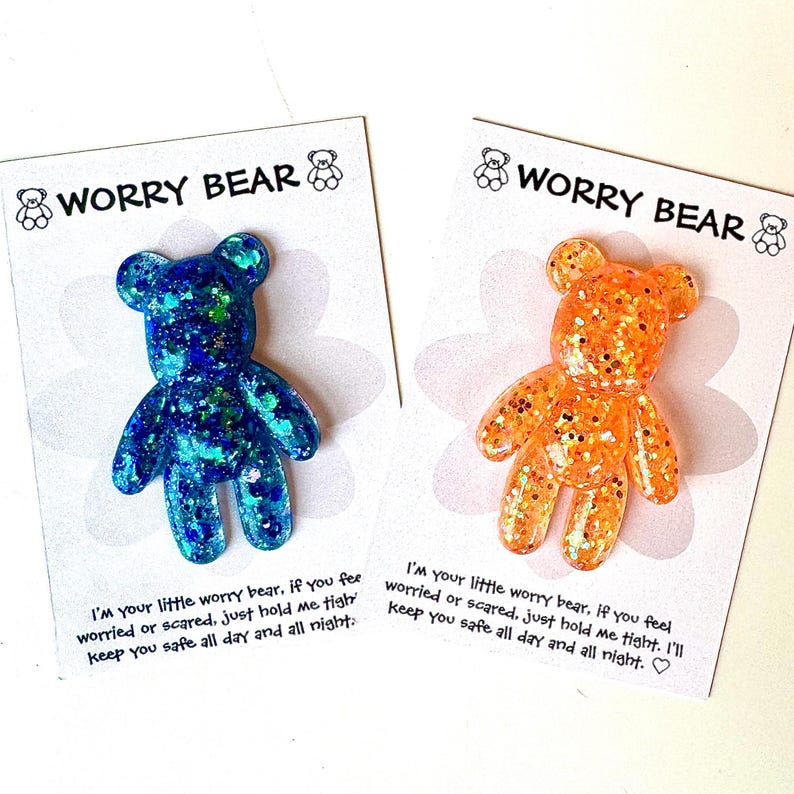Op de afbeelding: Twee glitterhars Worry Bear-bedels, &eacute;&eacute;n blauw en &eacute;&eacute;n oranje, op witte kaarten. Elke kaart heeft de tekst "Worry Bear" en een geruststellende boodschap. De beren zijn gevormd als teddyberen.
