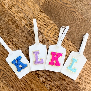 May include: Four white luggage tags with different colored felt letters. The letters are K, L, and K. The tags have a white strap for attaching to luggage.