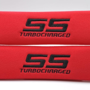 May include: Two red seat belt covers with black embroidered text. Each cover displays the number "55" above the word "TURBOCHARGED". The covers are made of a soft, textured material.