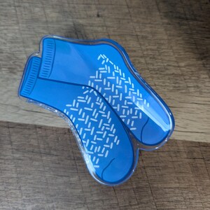 May include: A blue acrylic hair clip shaped like a pair of ankle socks. The clip features a vibrant blue color with a white, repeating dash pattern across the foot area and ribbed detailing at the ankle cuff, finished with a clear glossy coating.
