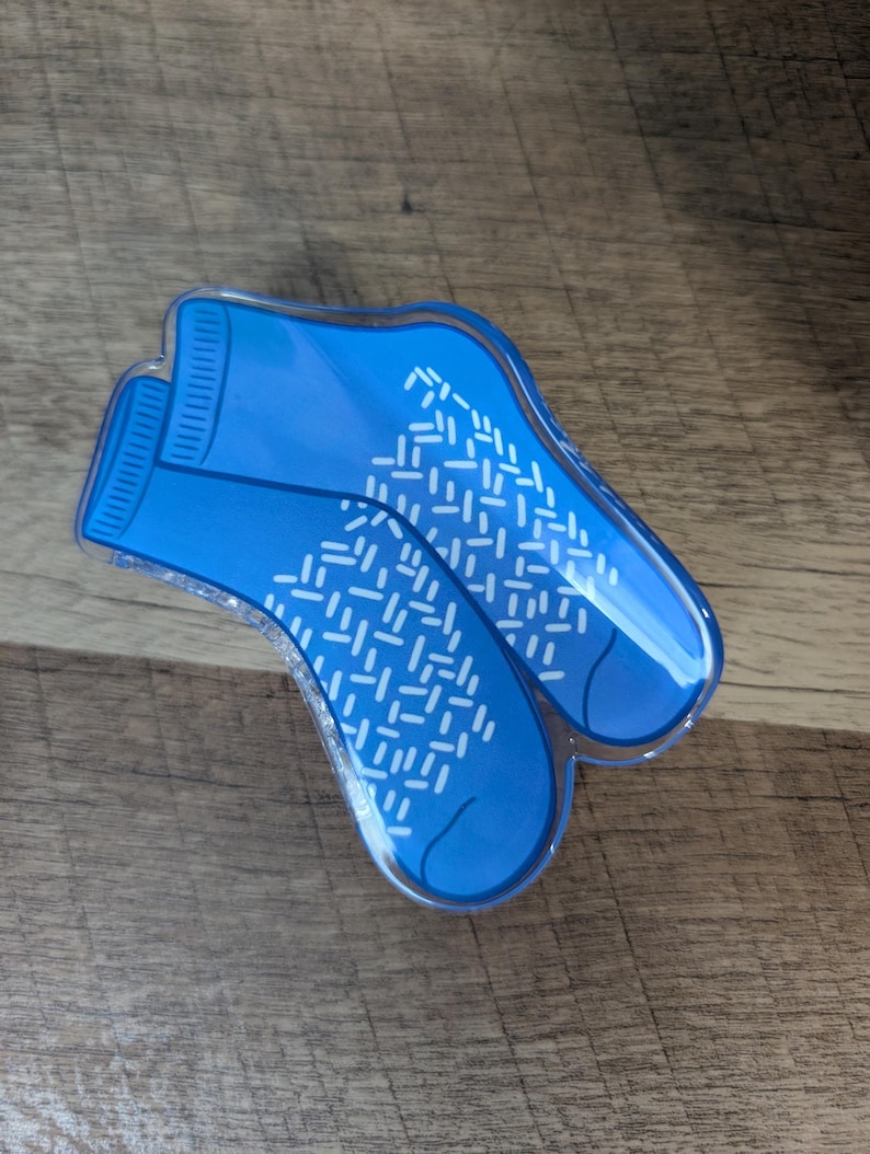 May include: A blue acrylic hair clip shaped like a pair of ankle socks. The clip features a vibrant blue color with a white, repeating dash pattern across the foot area and ribbed detailing at the ankle cuff, finished with a clear glossy coating.