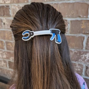 May include: A hair clip shaped like a reacher grabber tool. The clip features a white handle and shaft with a black and blue trigger mechanism and a black and blue claw at the end. The design is flat with a white outline, securing a section of hair.