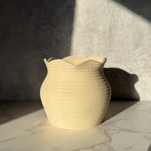 May include: A cream-colored, textured vase with a wavy rim. The vase has a cylindrical body with horizontal ridges. The background is a gray wall with a marble surface in the foreground. The lighting creates shadows.