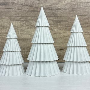 Puede incluir: Tres árboles de Navidad de papel blanco de diferentes tamaños, dispuestos sobre una superficie blanca. Los árboles están hechos de papel doblado y tienen un diseño en capas y escalonado.