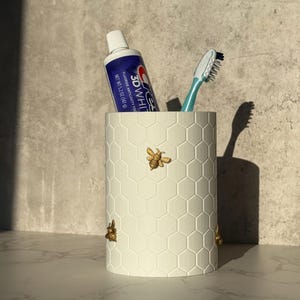 Könnte beinhalten: Ein weißer, zylindrischer Zahnbürstenhalter mit Wabenmuster und drei goldfarbenen Bienenverzierungen. Eine Tube Crest 3D White Zahnpasta und eine blaue Zahnbürste befinden sich im Inneren. Der Hintergrund ist ein neutrales Grau.