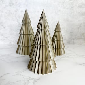 May include: Three decorative, tiered Christmas trees in varying sizes. The trees are a muted gold colour and have a layered, pleated design. The trees are arranged on a white marble surface.