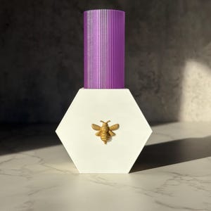 May include: A white, hexagonal bottle with a gold bee emblem and a ribbed, purple cap. The product is displayed on a white marble surface with a dark grey background.