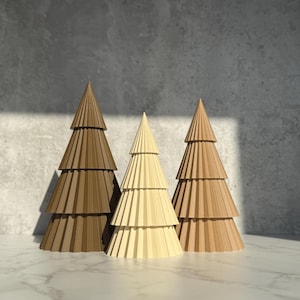 May include: Three decorative paper Christmas trees in shades of brown and beige. The trees have a layered, pleated design, with a pointed top. The trees are arranged on a white marble surface against a grey background.