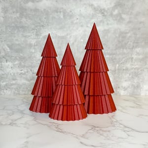 Könnte beinhalten: Drei dekorative rote Weihnachtsbäume mit einem geschichteten Design. Die Bäume sind in einer Reihe angeordnet, jeder mit einer spitzen Spitze und einem strukturierten, geriffelten Aussehen. Die Bäume stehen auf einer weißen Marmoroberfläche.