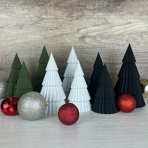 Könnte beinhalten: Ein Set aus sieben Papier-Weihnachtsbäumen in Grün, Weiß und Schwarz. Die Bäume sind in einer Reihe auf einer weißen Oberfläche angeordnet. Vor den Bäumen befinden sich auch rote und silberne Kugeln.