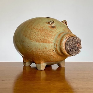 May include: A ceramic piggy bank with a cork stopper for the coin slot. The piggy bank is glazed in a light green and brown color with a speckled pattern.