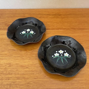 May include: Two black ceramic coasters with a wavy edge. Each coaster features a white flower design with green leaves.