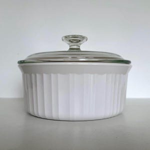May include: A white, round casserole dish with a clear glass lid and handle. The dish has vertical ridged detailing around the sides. The lid has a clear knob handle. The dish is on a white surface.