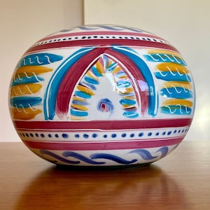 May include: A white ceramic sphere with a colourful painted design. The design features blue, yellow, red and green stripes and swirls.
