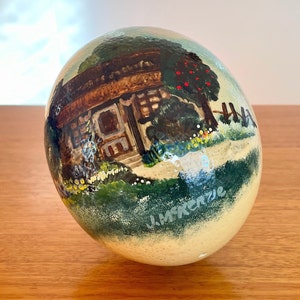 以下が含まれることがあります： 手描きのダチョウの卵で、赤い屋根の小さな小屋と赤いリンゴのついた緑の木という幻想的なシーンが描かれています。卵は薄いベージュの背景に緑の草と花が描かれています。アーティストの署名「J. McKenzie」は卵の右下にあります。