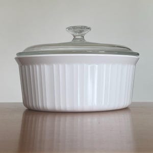 May include: White ceramic baking dish with a glass lid. The dish has a fluted design and is ideal for serving casseroles, pasta dishes, or other baked goods.