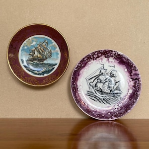 May include: Two decorative plates with a nautical theme. One plate features a burgundy background with gold accents and a black and white illustration of a sailing ship. The other plate has a purple background with a white centre and a black and white illustration of a sailing ship.