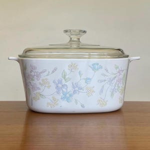 May include: White oval casserole dish with a floral design in shades of blue, purple, and green. The dish has a clear glass lid with a knob.